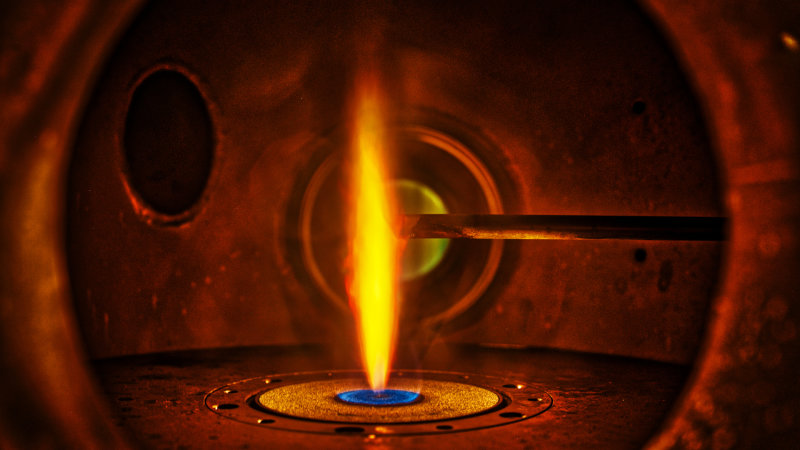 Blick auf die Mitte eines Synthesereaktors, in dem Nanopartikel innerhalb einer Sprayflamme entstehen und untersucht werden – zum Beispiel für die Entwicklung neuer Katalysatormaterialien. Die in die Flamme ragende Sonde zur Probenentnahme hält Temperaturen bis zu 2.600°C stand.