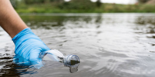 Foto: Eine Hand mit blauem Einmal-Handschuh hält eine Wasserflasche in ein Gewässer in der Natur.