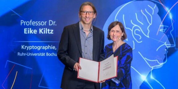 Ein Mann und eine Frau, die vor einem blauen Hintergrund stehen. Der Mann hält eine Urkunde.