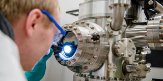 Foto: Nahaufnahme einer blonden Person mit Brille schaut sich eine Sputteranlage mit einer Taschenlampe an.