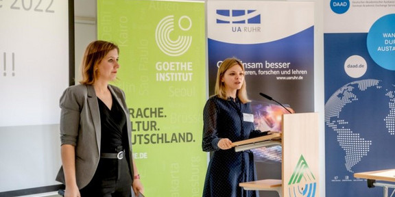 Zwei Frauen stehen vor Aufstellern der UA-Ruhr. Eine Frau steht an einem Pult mit Mikrofon und hält eine Rede.