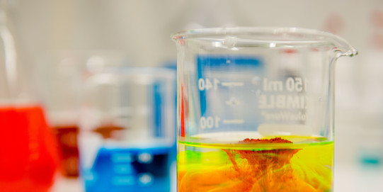 Close-up of a laboratory vessel in which a red and a yellow liquid are mixed, in the background other vessels with colored liquids