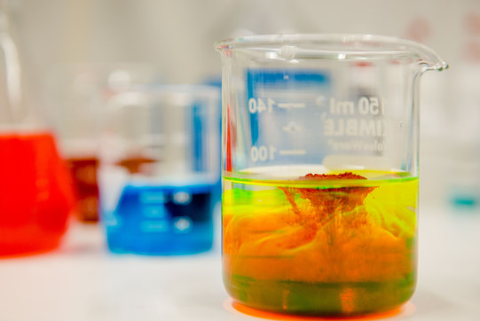 Close-up of a laboratory vessel in which a red and a yellow liquid are mixed, in the background other vessels with colored liquids
