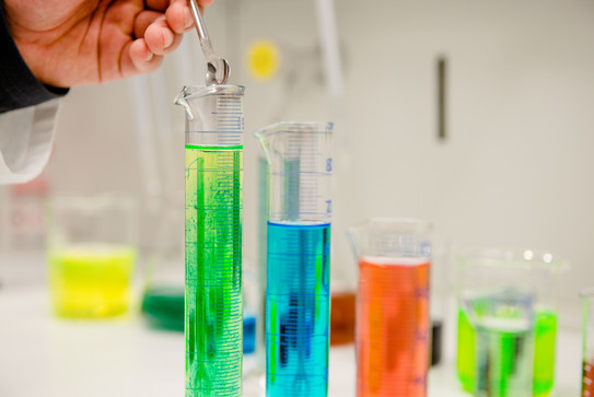 Eine Reihe von Reagenzgläsern mit verschiedenfarbigen Flüssigkeiten, eine Hand füllt etwas mit einem Löffel in das Glas ganz links