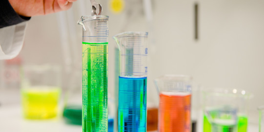 Eine Reihe von Reagenzgläsern mit verschiedenfarbigen Flüssigkeiten, eine Hand füllt etwas mit einem Löffel in das Glas ganz links