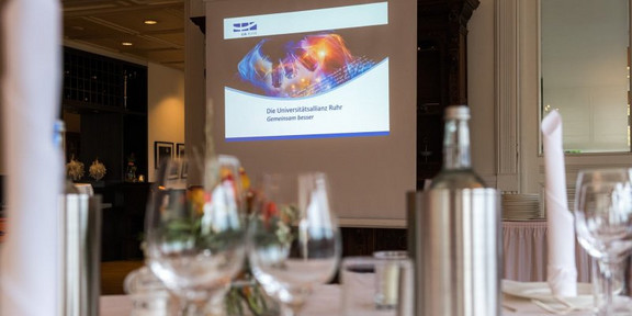 Das Bild zeigt einen mit weißer Tischdecke gedeckten Tisch. Im Hintergrund ist eine Präsentation der Universitätsallianz Ruhr zu sehen mit dem Motto "Gemeinsam besser".