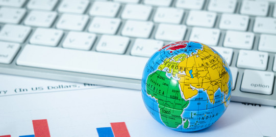 Ein kleiner bunter Globus liegt vor einer modernen Tastatur und auf einem Ausdruck mit Säulendiagrammen