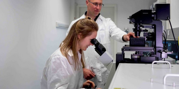 Eine Wissenschaftlerin, die in ein Mikroskop schaut. Hinter ihr steht ein weiterer Wissenschaftler. 