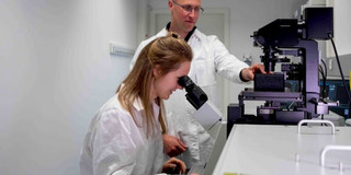 Eine Wissenschaftlerin, die in ein Mikroskop schaut. Hinter ihr steht ein weiterer Wissenschaftler. 
