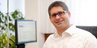 Das Bild zeigt Prof. Sven Rahmann am Schreibtisch sitzend. Er trägt eine Brille mit schwarzem Rahmen und ein langärmeliges weißes Hemd.