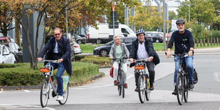 Drei Männer und eine Frau, die mit Fahrrädern über einen Parkplatz an der TU Dortmund fahren.