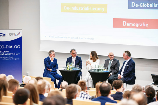 Fünf Personen sitzen in einer Talkrunde vor einer großen Leinwand, im Vordergrund Hinterköpfe des Publikums.