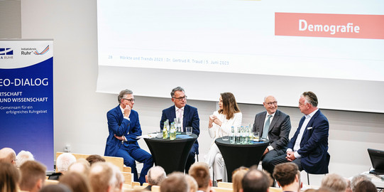 Fünf Personen sitzen in einer Talkrunde vor einer großen Leinwand, im Vordergrund Hinterköpfe des Publikums.