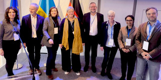 A group of nine people stand next to each other, smiling and wearing name badges. Flags can be seen in the background.