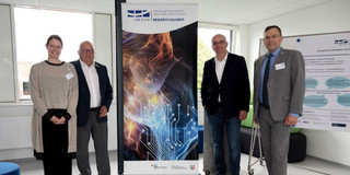 Four people standing next to a poster of the Research Alliance of the UA Ruhr.