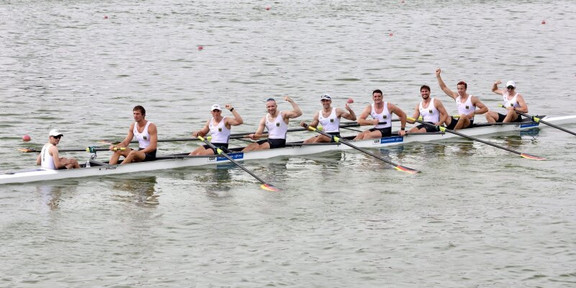 Foto: Ein Ruderboot mit acht Ruderern fährt über Wasser. Einige der Ruderer recken einn Arm triumphierend in die Luft.