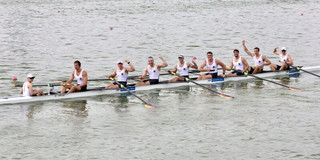 Foto: Ein Ruderboot mit acht Ruderern fährt über Wasser. Einige der Ruderer recken einn Arm triumphierend in die Luft.