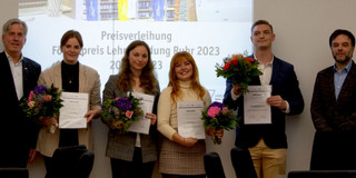 Foto: Eine Gruppe Studierender hält Blumen und Urkunden in den Händen. Hinter ihnen sieht man eine Projektion an der Wand. Neben ihnen stehen zwei Männer.