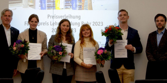 Foto: Eine Gruppe Studierender hält Blumen und Urkunden in den Händen. Hinter ihnen sieht man eine Projektion an der Wand. Neben ihnen stehen zwei Männer.