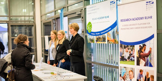 Das Bild zeigt einen Informationsstand der Research Academy Ruhr. Auf dem Bild sind Banner, eine Theke mit Flyern sowie Personen zu sehen.