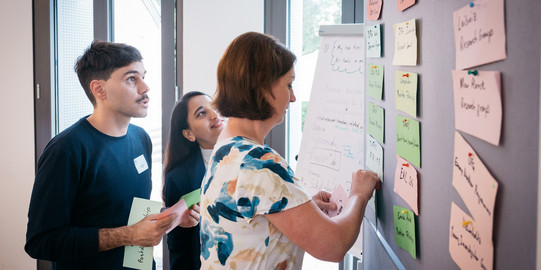 Three people are standing in front of a wall, pinning written cards on it.