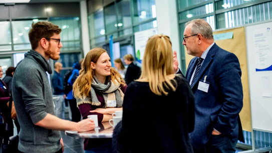 Four people of different ages stand at a high table in a foyer and discuss