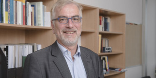Porträtfoto: Prof. Johannes Weyer, ein Mann mit grauen Bart und blauer Brille, steht in einem Büro vor einem Bücherregal und lächelt in die Kamera.