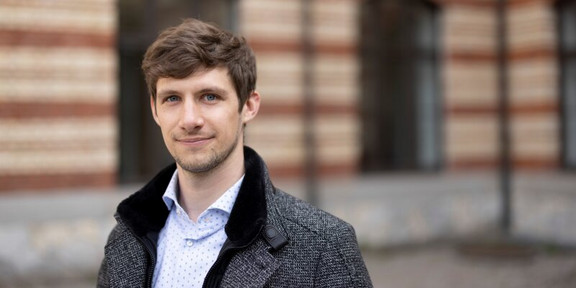 Photo: Portrait photo of a man with brown hair in a gray coat. A blurred building can be seen in the background.