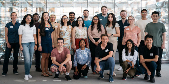 Foto: Eine Gruppenfoto internationaler Nachwuchswissenschaftler*innen vor einer Glaswand.