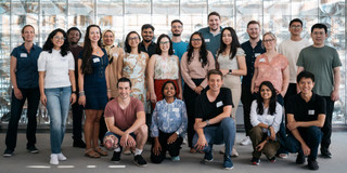 Foto: Eine Gruppenfoto internationaler Nachwuchswissenschaftler*innen vor einer Glaswand.