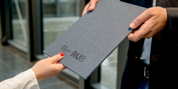 Das Bild zeigt eine Mappe mit der Aufschrift "RUB" und wird von zwei unterschiedlichen Personen gehalten.