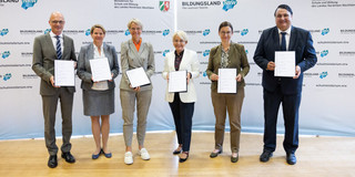 Gruppenfoto: Sechs Personen in formaler Kleidung stehen vor einem Banner mit der Aufschrift Bildungsland NRW und halten Urkunden in den Händen.