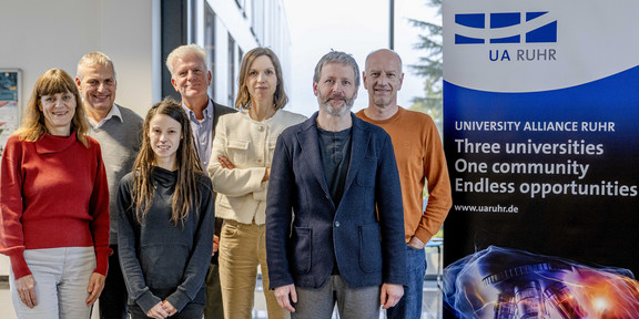 Foto: Ein Gruppenfoto vom UA Ruhr Rat für Ethik und Verantwortung neben einem Aufsteller der UA Ruhr. Auf dem Aufsteller steht: "University Alliance Ruhr. Three universities, one community, endless opportunities"