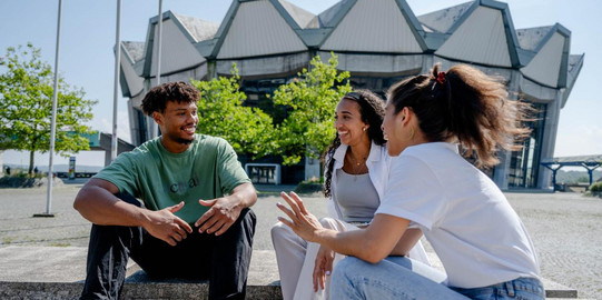 Two female and one male student sitting in a casual conversation with the Audimax of the Ruhr University Bochum in the background.