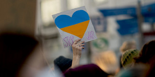 Foto: Eine Hand hält bei einem Protest ein Plakat hoch, auf dem ein Herz in den Farben der Ukraine abgebildet ist. Neben dem Herz steht "Stop War".