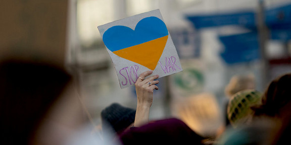 Foto: Eine Hand hält bei einem Protest ein Plakat hoch, auf dem ein Herz in den Farben der Ukraine abgebildet ist. Neben dem Herz steht "Stop War".