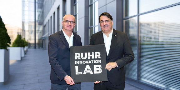 Zwei Rektoren halten ein schwarzes Schild mit der Aufschrift "Ruhr Innovation Lab" hoch.